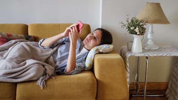 Young brunette woman feel sick and rest at home during the day under the plaid on the sofa scrolling her smartphone