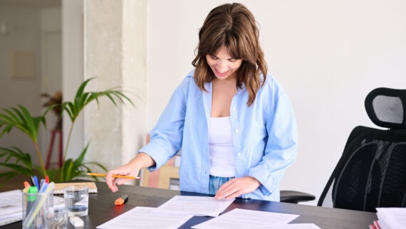 ChatGPT  A young woman is standing at a desk, looking down and smiling as she reviews documents spread out in front of her. She is wearing a light blue shirt over a white top and jeans. The workspace is well-organised, with office supplies, a glass of water, and a potted plant nearby. The background is bright and minimalist, creating a professional and focused atmosphere. The scene suggests she is engaged in detailed work or preparing materials, reflecting a positive and productive work environment.