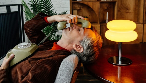 A man with grey hair is leaning back in a chair, holding an old-fashioned rotary phone to his face. He appears to be frustrated or overwhelmed. The setting is a vintage-style room with warm lighting, a wooden cabinet in the background, a potted plant, and a yellow table lamp on a round table next to him. The scene evokes a sense of nostalgia and stress.