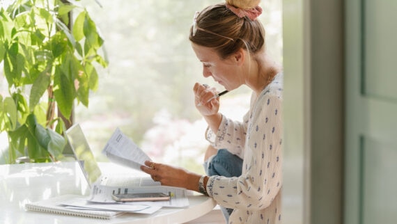 woman concentrating in a bright home office, looking at papers by a laptop and mobile phone