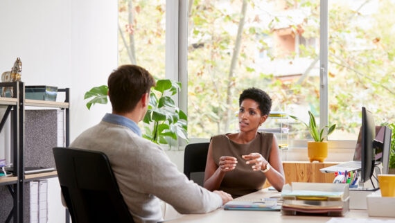 A woman and a man are seated at a desk in an office, engaged in a serious conversation. The woman is speaking and gesturing with her hands, while the man listens attentively. The office is well-lit with large windows and green plants, creating a pleasant atmosphere.