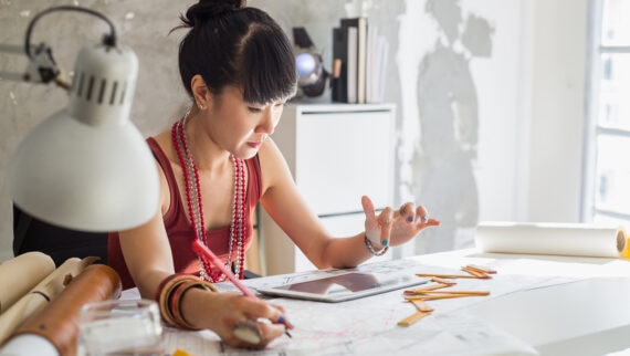 A focused female designer examines architectural plans at her brightly lit studio desk, surrounded by various design tools and with a digital tablet at hand.