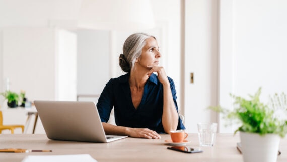 A woman with grey hair, tied back neatly, is seated at a desk in a bright, modern office. She is wearing a dark blue blouse and appears to be deep in thought, looking out of the window. In front of her is a laptop, an orange coffee cup, a glass of water, and a few other office supplies. The background features a clean and minimalist design, with a few green plants adding a touch of nature to the workspace. The scene conveys a contemplative and serene atmosphere.