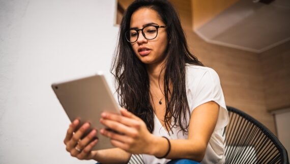 A concentrated young woman wearing glasses, using a tablet in a modern, well-lit indoor setting.