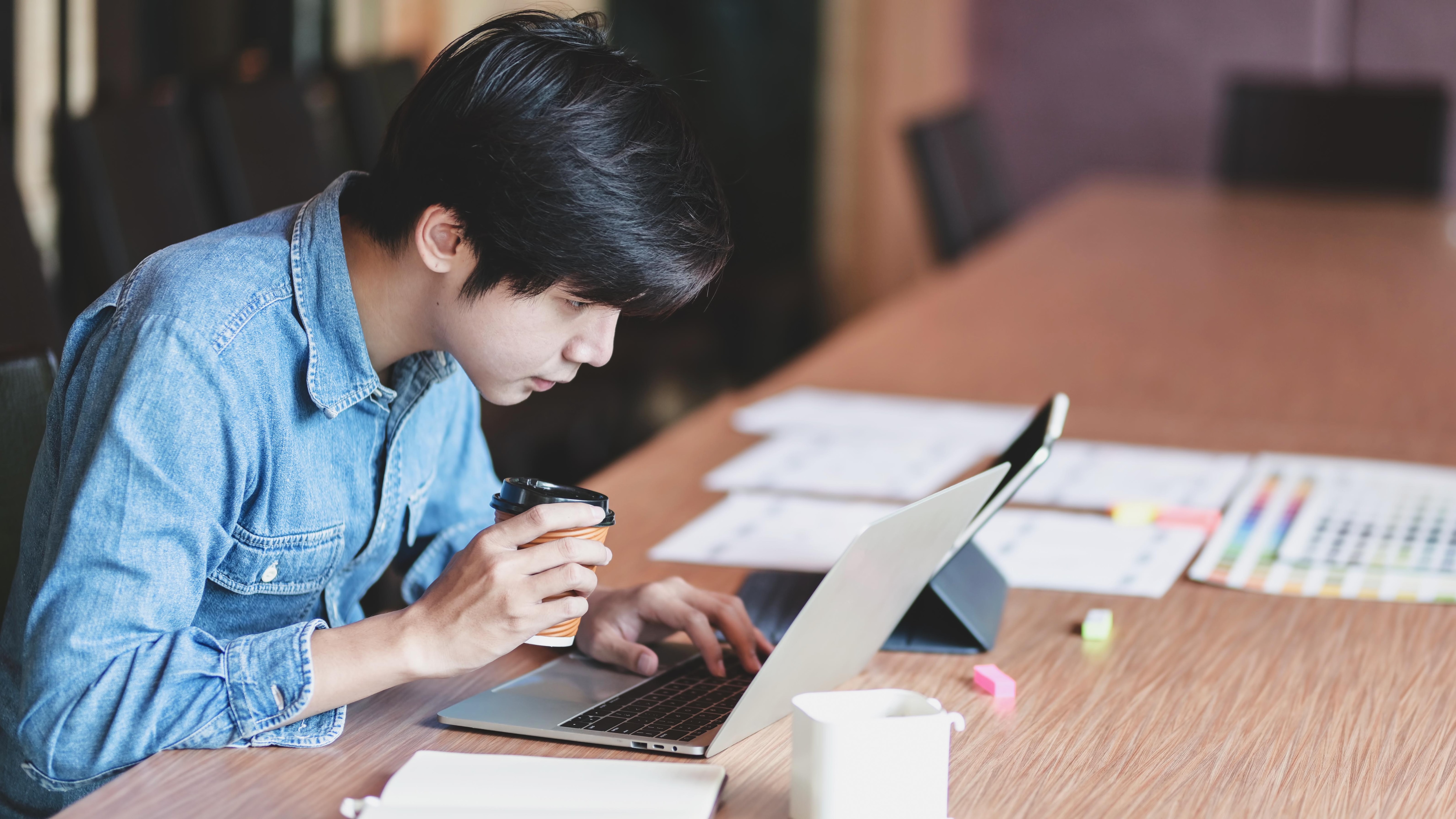 A focused young man with black hair, wearing a denim jacket, drinking coffee while working on his laptop at a busy desk strewn with papers and sticky notes.