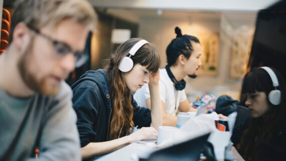 Three people working in a desk. The woman in middle is wearing headphones and staring at her laptop screen.