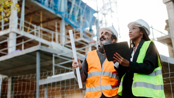 Two construction workers, a man and a woman, are at a building site. Both are wearing safety vests and hard hats. The man, holding a rolled-up blueprint, is pointing towards something on the construction site, while the woman holds a tablet, possibly reviewing plans or data. The background shows a partially constructed building with scaffolding. The scene suggests they are discussing the project's progress or specific construction details, indicating a collaborative and professional interaction in a construction setting.