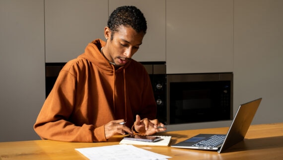 A young man in an orange hoodie is seated at a wooden table, deeply focused on his work. He is using a smartphone and writing in a notebook, with a laptop open nearby. Papers with handwritten notes are spread out on the table, indicating he is engaged in detailed tasks. The background features a modern kitchen setting with built-in appliances, providing a warm and contemporary atmosphere. The scene conveys a sense of concentration and productivity in a home office environment.