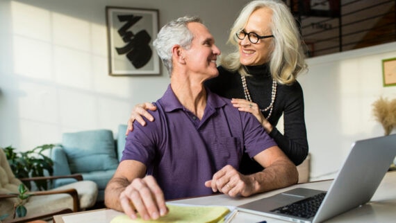 An elderly couple smiling at each other while working at a desk with a laptop and documents in a bright living room.