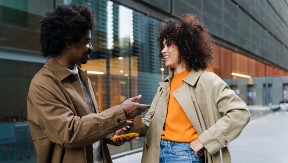 A man and a woman are standing outside a modern building, engaged in a lively conversation. Both are wearing light brown coats; the woman is also wearing an orange top and jeans. The man is holding a yellow phone in one hand while gesturing with the other. They appear to be enjoying the conversation, smiling and maintaining eye contact. The setting is urban, with glass windows and a sleek architectural background.