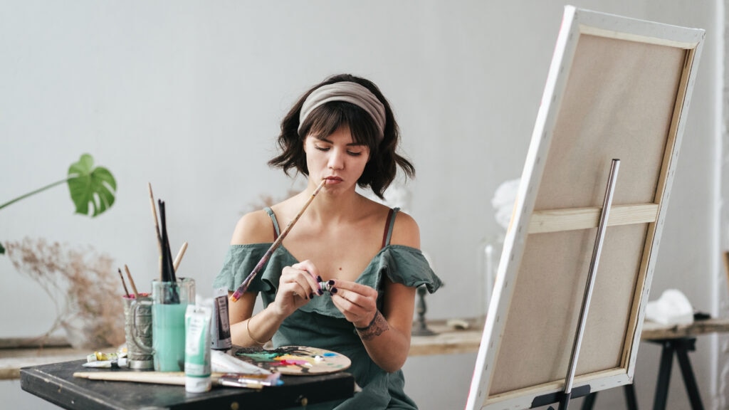 Woman sitting in her studio painting on a canvas. She is holding the paint brush with her mouth whilst she is opening a colour tube.