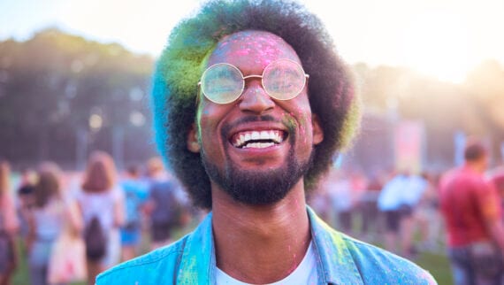 A man wearing glasses smiling at the camera. He is covered in colourful dust.