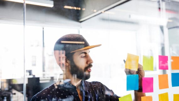 A portrait of man wearing a cap, and standing in front of a glass wall full of coloured sticky notes.