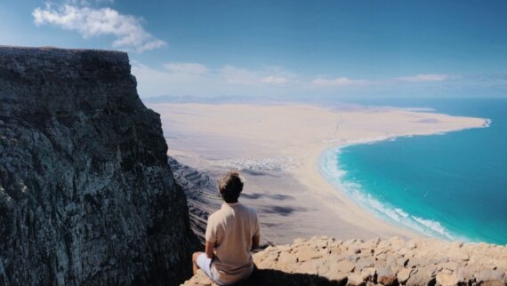 A man sitting on top of a cliff overlooking the ocean.