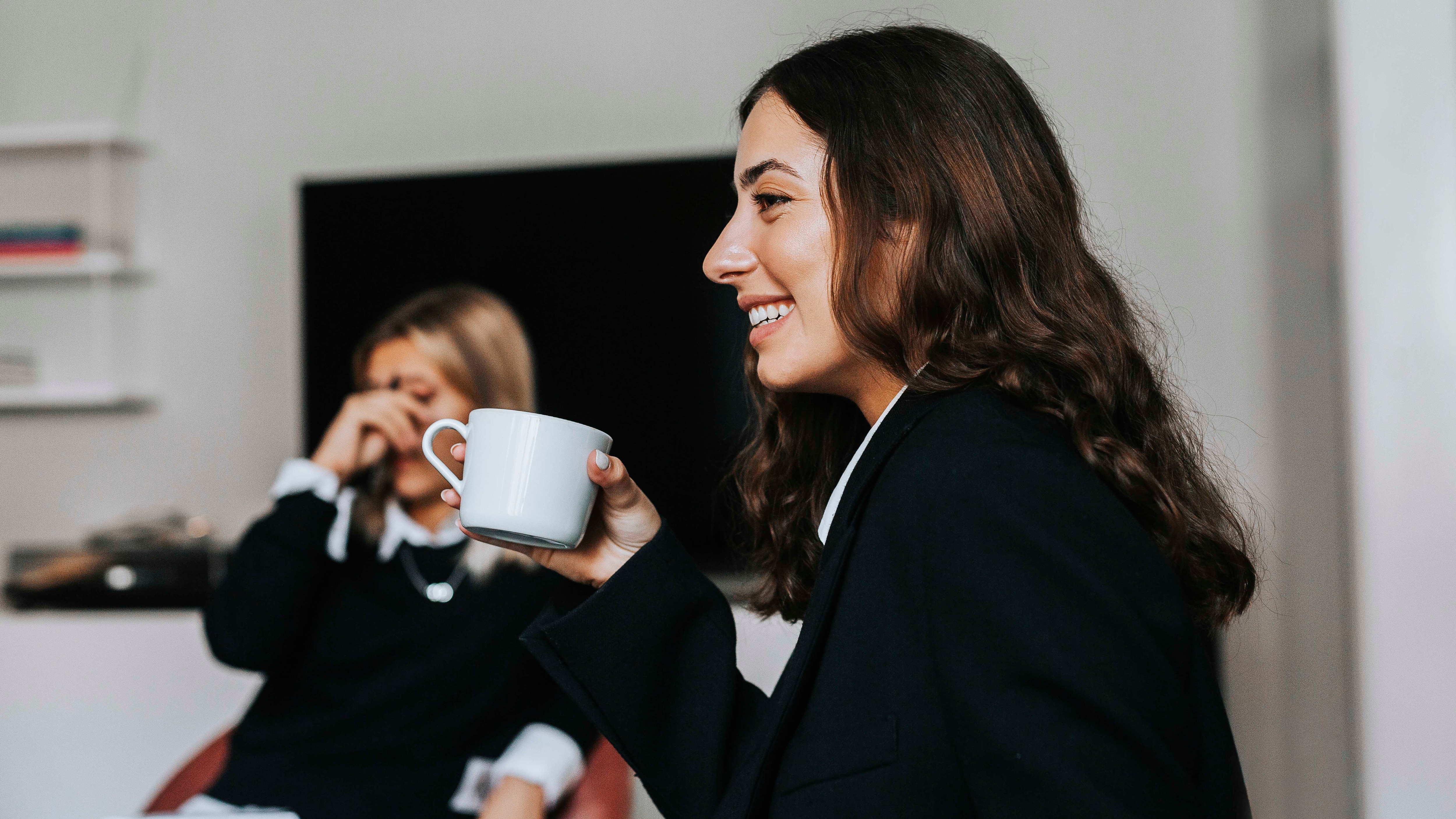A woman with long brown hair is smiling and holding a cup of coffee. She is wearing a black blazer and appears to be enjoying a conversation. In the background, another woman is seated, also dressed in dark clothing, with her hand resting on her face, appearing relaxed. The setting suggests a casual and comfortable environment, possibly a lounge or office break room.