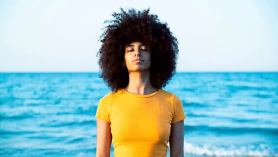 Young Afro woman with eyes closed standing at beach and taking a deep breath in.