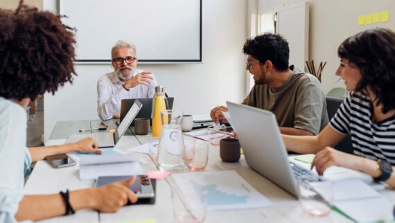 A group of four people is seated around a table in a meeting room, engaged in discussion. An older man with grey hair and glasses is speaking, while the other three, consisting of two women and a man, listen attentively. They have laptops, tablets, and documents spread out on the table. The background features a whiteboard or screen, and the setting appears to be collaborative and professional.