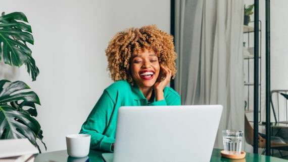 A woman with curly hair is sitting at a desk, smiling brightly as she looks at her laptop. She is wearing a green top and appears to be engaged in a virtual meeting or conversation. On the desk, there is a white mug and a glass of water, along with some documents. The background includes a large leafy plant and a modern, well-lit room, creating a cheerful and professional environment.