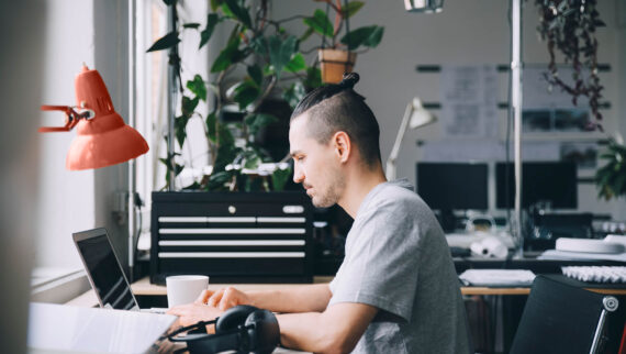 Young man in a gray T-shirt sits at a desk and works with a laptop.