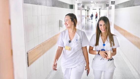 Two women, dressed in white medical uniforms, are walking down a hospital corridor, smiling and chatting with each other. Both are wearing identification badges, and one is holding a small notebook and a pair of scissors. The corridor is brightly lit and clean, with tiled walls and a handrail running along the side. The atmosphere is friendly and professional, reflecting a positive work environment in a healthcare setting.