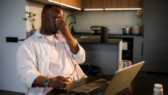 The man took off his glasses and rubs his eyes tired of working on a laptop. An open laptop is on the kitchen table at home. Daylight