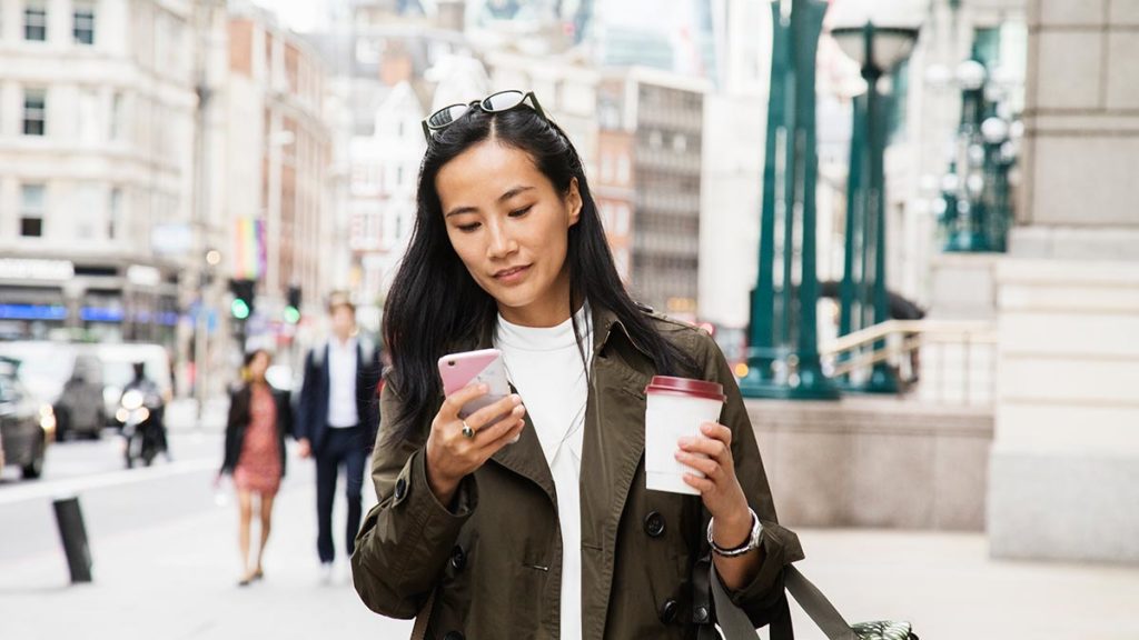 A woman walking down the street looking at her phone, with a coffee in her hand.