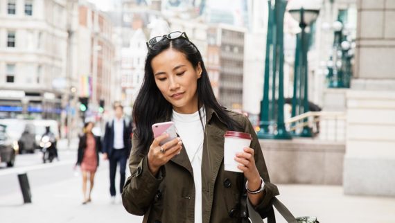 A woman walking down the street looking at her phone, with a coffee in her hand.