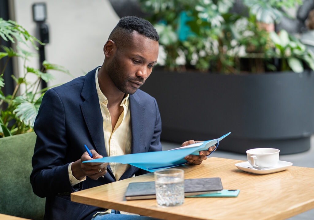 A smartly dressed man sits at a wooden table in a modern indoor setting, reviewing documents held in a blue folder. He appears focused, with a pen in hand. On the table are a cup of coffee, a glass of water, a notebook, and a smartphone. Lush green plants can be seen in the background.
