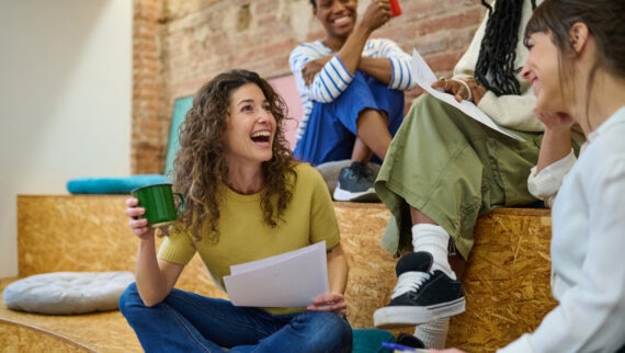 A group of colleagues gathered on tiered wooden seating in a casual workspace. One woman in a green sweater sits cross-legged on the floor, holding a green mug and a sheet of paper while laughing. Two others sit above her—one in striped top holding a red mug, another in light clothing smiling—each with papers in hand, engaged in a relaxed discussion.