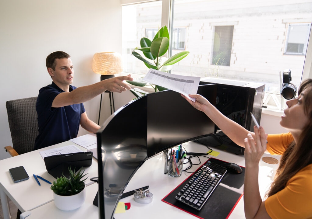 A man sitting at a desk in an office, reaching out to take a printed document from a woman seated across from him. The workspace features dual curved monitors, a keyboard, various stationery items, a small potted plant, and a large window letting in natural light. The woman holds a pen and appears to be explaining something.
