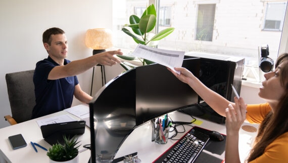 A man sitting at a desk in an office, reaching out to take a printed document from a woman seated across from him. The workspace features dual curved monitors, a keyboard, various stationery items, a small potted plant, and a large window letting in natural light. The woman holds a pen and appears to be explaining something.