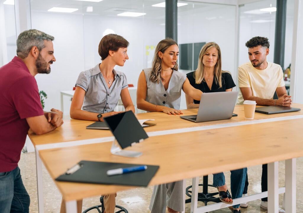 A group of five colleagues gathered around a high desk in a modern office, engaged in discussion. Two women are seated, one of whom is pointing at a laptop screen, while the others stand or lean in attentively. Notebooks, a tablet, and a takeaway coffee cup are on the desk. The office space features glass walls and a bright, open layout.