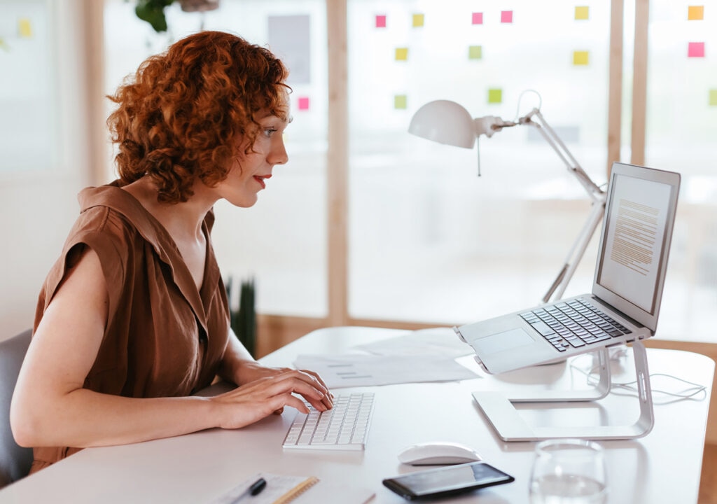 A woman with curly red hair working at a desk in a bright, modern office. She is typing on a wireless keyboard, with a laptop placed on a stand in front of her. The desk also holds a mouse, tablet, notebook, and a glass of water. Colourful sticky notes are visible on the glass wall in the background.