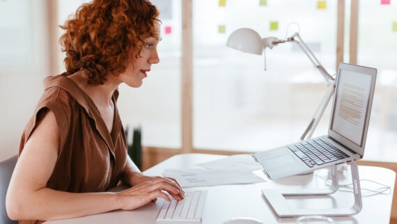 A woman with curly red hair working at a desk in a bright, modern office. She is typing on a wireless keyboard, with a laptop placed on a stand in front of her. The desk also holds a mouse, tablet, notebook, and a glass of water. Colourful sticky notes are visible on the glass wall in the background.