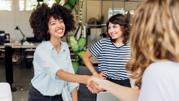 A smiling woman confidently shaking hands with an interviewer in a modern office, with a colleague observing in the background—illustrating a positive competency-based interview setting