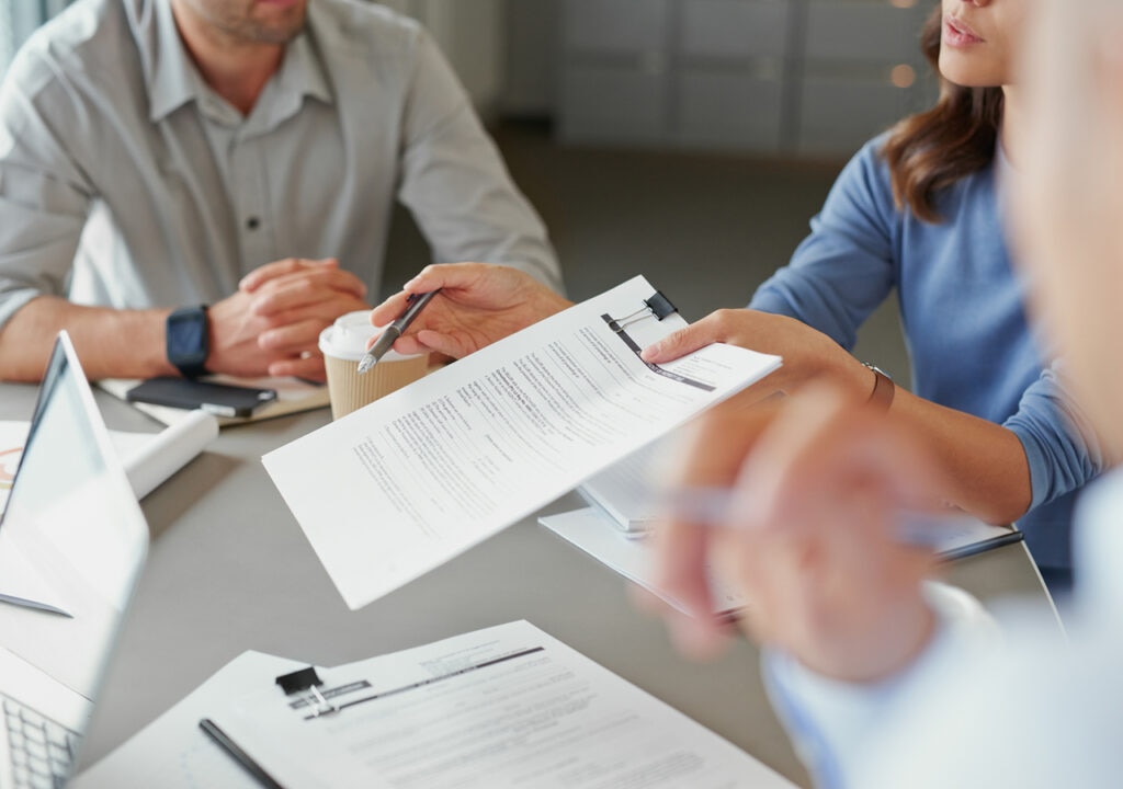 Close-up of a business meeting, with one person holding and pointing at a printed document clipped to a folder. Other participants are seated around the table, with laptops, pens, and takeaway coffee cups visible. The focus is on the discussion of the document, with hands and paperwork in the foreground.