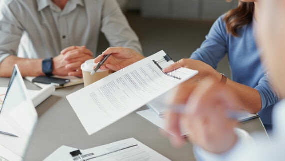 Close-up of a business meeting, with one person holding and pointing at a printed document clipped to a folder. Other participants are seated around the table, with laptops, pens, and takeaway coffee cups visible. The focus is on the discussion of the document, with hands and paperwork in the foreground.