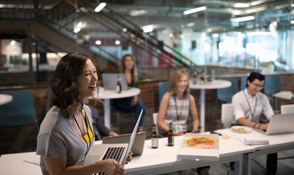 An image of a woman holding a laptop in a modern office, with colleagues eating pizza in the background.