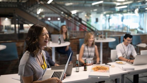 An image of a woman holding a laptop in a modern office, with colleagues eating pizza in the background.