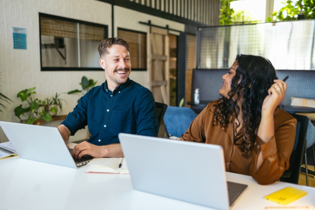 Two cheerful coworkers talking and sharing ideas in the office while working with laptops, highlighting the value of an engaged, motivated, and loyal workforce
