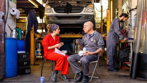 An image of a younger worker socialising with an older worker during their lunchbreak, outside a workshop.