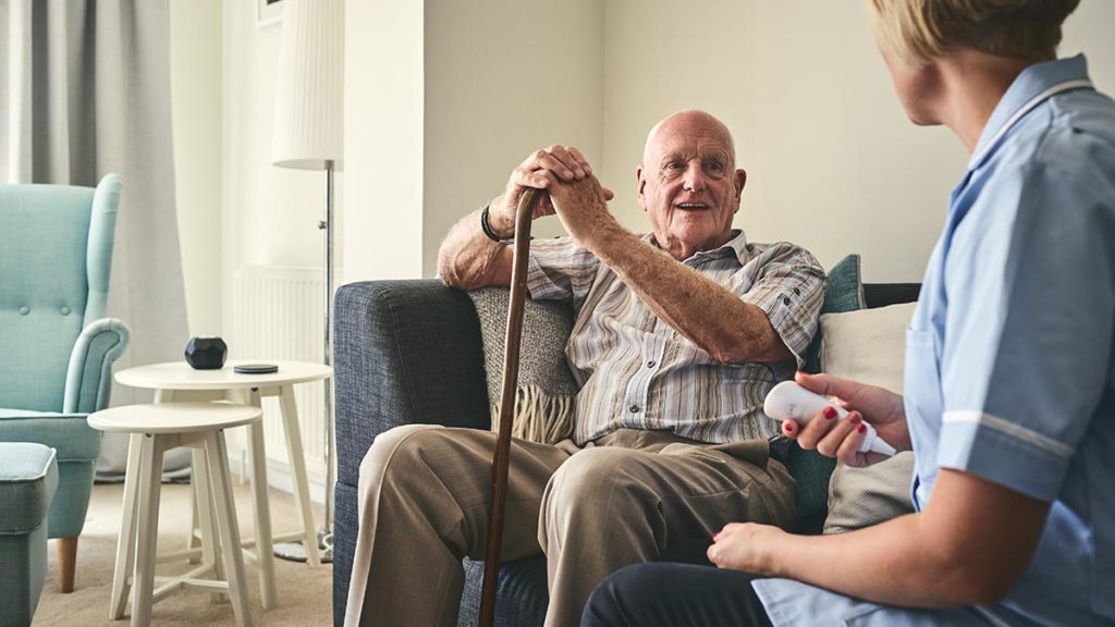 An image of an elderly man holding a walking stick, alongside a nurse who is caring for him.