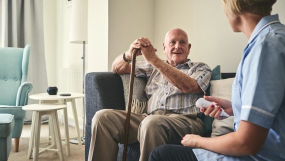 An image of an elderly man holding a walking stick, alongside a nurse who is caring for him.