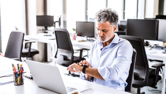 A man in an office surrounded by empty desks, checking his watch.