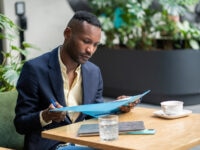 A smartly dressed man sits at a wooden table in a modern indoor setting, reviewing documents held in a blue folder. He appears focused, with a pen in hand. On the table are a cup of coffee, a glass of water, a notebook, and a smartphone. Lush green plants can be seen in the background.