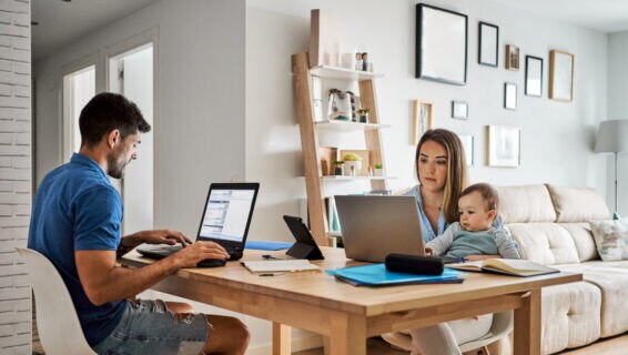 A man and a woman work on laptops at a wooden dining table in a modern living room. The woman is holding a baby on her lap while typing, and the man is focused on his screen. Shelves, framed pictures, and a beige sofa are in the background.