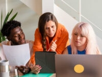 Three colleagues – a Black man holding a document, a brunette woman pointing at a tablet with a stylus, and a blonde woman viewing a laptop – collaborating at a desk in a modern office.