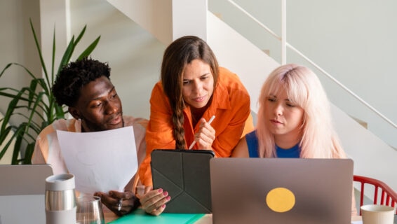 Three colleagues – a Black man holding a document, a brunette woman pointing at a tablet with a stylus, and a blonde woman viewing a laptop – collaborating at a desk in a modern office.