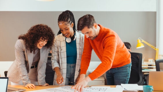 Three colleagues collaborate over a desk, smiling as they review printed plans in a modern office.