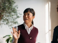 Two colleagues engaged in a discussion, one holding a pen and gesturing while speaking, with the other smiling and listening in a bright office setting.